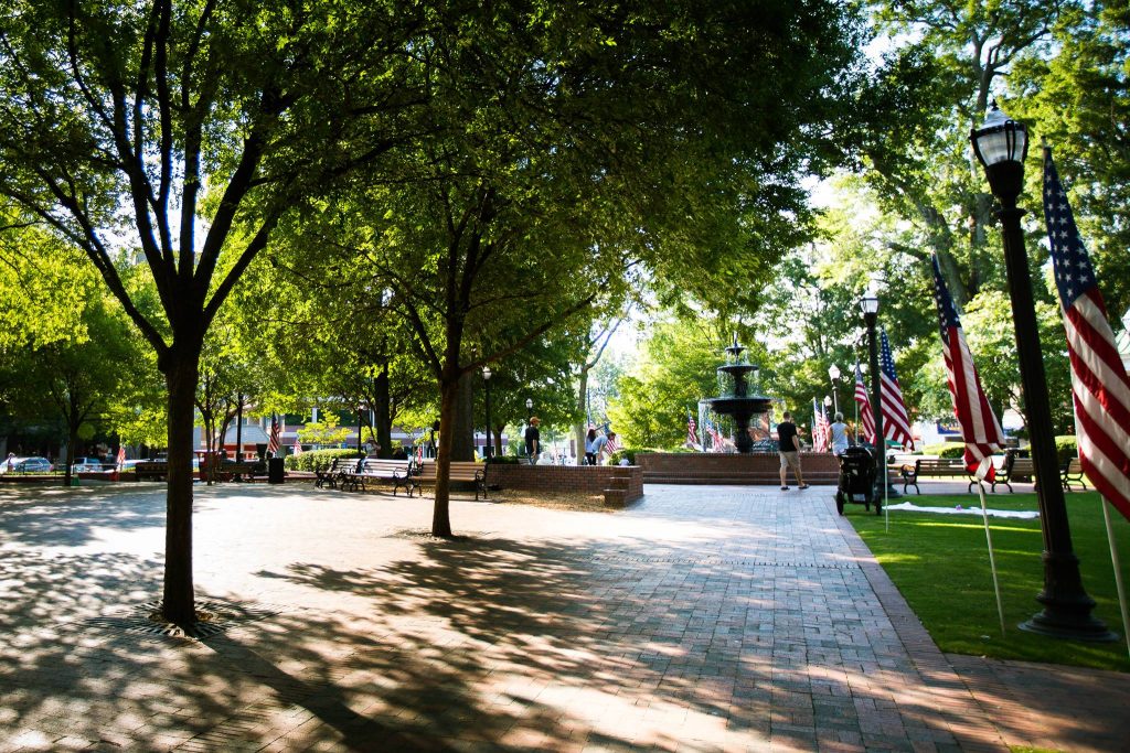 A vibrant park with a fountain surrounded by lush green trees, benches, and American flags, capturing a sunny day atmosphere.